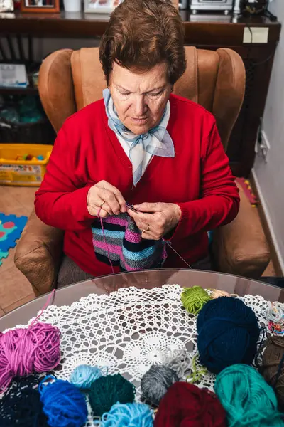 portrait of senior woman and movement of her hands sewing with needles and colored wool on a sofa at home