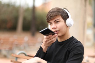 Kulaklıklı, cep telefonuyla dikte eden genç bir erkek parktaki bankta oturuyor.