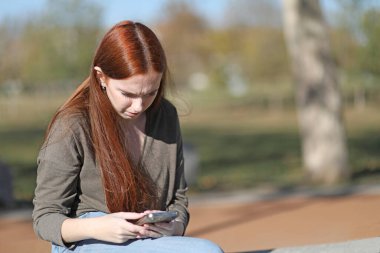 Parkta tek başına oturan bir kadın cep telefonuna bakıyor.