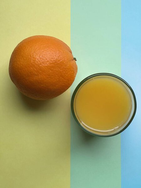 Апельсиновый сок и оранжевый на ярком фоне, состоящем из трех цветов: желтого, светло-зеленого и синего. Вид сверху крупным планом.