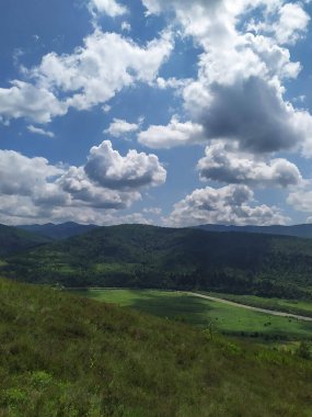 Gökyüzündeki bulutların altında yeşil dağlar görebilirsin.