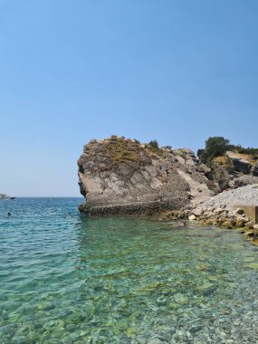 Kocaman bir kayayla deniz manzarası. Mavi güneşli, açık gökyüzü. Taşta çalılıklar var. Açık su alanı. Yaz tatili. Dikey