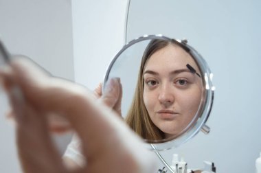 Kişisel bakım, kendi kendine bakım, eldivenli beyaz kadın, yüzünü makyaj, sabah rutininde aynaya bakarak kaşlarını fırçalama. Hafif arkaplan. Dizinin bir parçası.