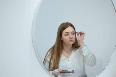 Genç beyaz bir kadın aynaya bakıyor, elinde köpükle kaşlarını temizlemek için bir fırça tutuyor. Ev konseptinde kendi kendini yetiştiren güzellik salonu. Bencillik. Mesaj için yer var. Dizinin bir parçası.