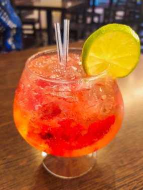 Bir bardak kırmızı meyve suyu cini ve tonik, limon dilimiyle süslenmiş, ahşap bir masada, bir içki barı atmosferinde.