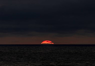 Gün sona ererken güneş yavaşça engin okyanusa batıyor ve batarken sıcak renkleriyle bulutlu gökyüzünü boyuyor.