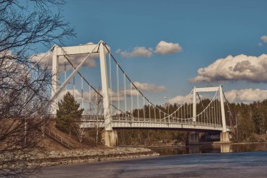Bereketli yeşillikler ve kabarık bulutlarla çevrili sakin bir nehrin üzerindeki asma köprü manzarası.