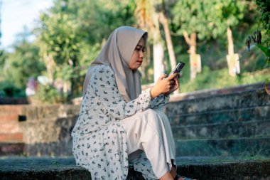 Modern tesettür giyen Asyalı kadın parkta akıllı telefonunu kullanarak sosyal medyada oynuyor - Asyalı kadın