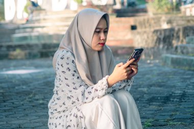 Modern tesettür giyen Asyalı kadın parkta akıllı telefonunu kullanarak sosyal medyada oynuyor - Asyalı kadın