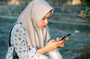 Modern tesettür giyen Asyalı kadın parkta akıllı telefonunu kullanarak sosyal medyada oynuyor - Asyalı kadın