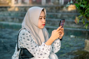 Modern tesettür giyen Asyalı kadın parkta akıllı telefonunu kullanarak sosyal medyada oynuyor - Asyalı kadın