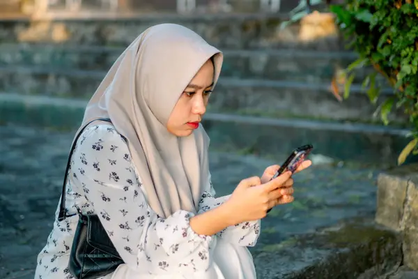 Stock image Asian woman wearing modern hijab playing social media using her smartphone in the park - Asian woman