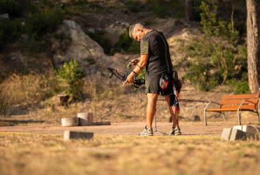 Bir adam elinde ok ve yayla bir tarlada duruyor. Siyah bir gömlek ve şort giyiyor.