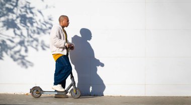 Şehirde beyaz duvarın yanında elektrikli scooter kullanan genç bir adam.