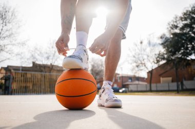 Basketbol ayakkabılarını bağlayan genç bir adam topunu ayaklarının arasına almış, açık bir sahada dikiliyor.