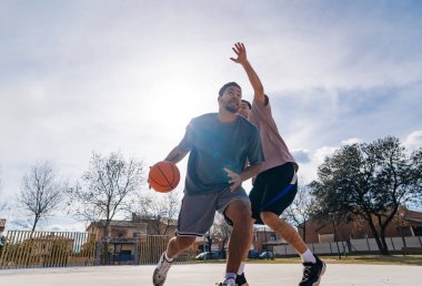 Güneşli bir günde, açık hava sahasında arkadaşça ama rekabetçi bir basketbol maçının keyfini çıkaran iki erkek arkadaş.