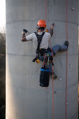 Endüstriyel tırmanışçı beton bacaya güvenlik ekipmanı ile matkap yerleştiriyor.