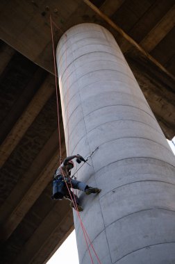 Endüstriyel tırmanıcı, köprü sütunu üzerinde çalışırken bir matkap kullanarak, yükseklerde güvenliği sağlıyordu.