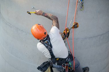 İp erişim teknisyeni beton bir duvarda iplere asılırken bir demir atma noktasını çekiçliyor.