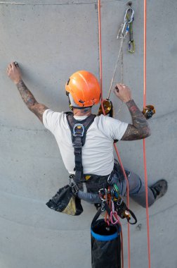Endüstriyel tırmanışçı. Dövmeleri ve kaskı iplere asılı ve beton duvarları denetliyor.