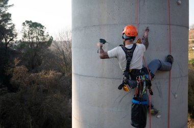 Endüstriyel tırmanıcı, beton baca üzerinde çalışırken ip erişim teknikleri kullanarak matkap kullanıyor.