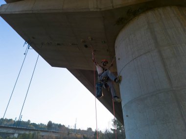 İp erişim teknisyeni tırmanma teçhizatı kullanarak beton köprü direğini inceliyor.