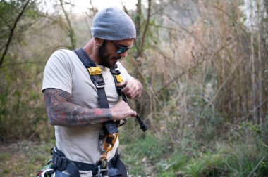 Tırmanışçı emniyet kemerini ayarlıyor ve ağaçlık bir alana tırmanmaya hazırlanıyor.