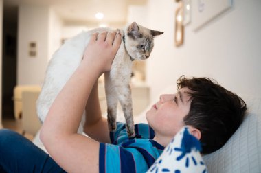 Çocuk kanepede uzanıyor, kedisini yukarıda tutuyor, birbirine bakıyor.