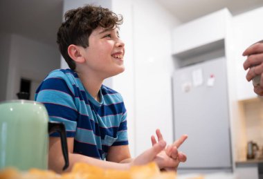 Gülümseyen çocuk modern bir mutfakta yemek yerken annesiyle sohbet ediyor.