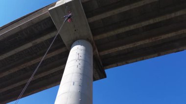 Profesyonel işçi iplerden sarkıyor, mavi gökyüzüne karşı beton köprü sütunlarını inceliyor.