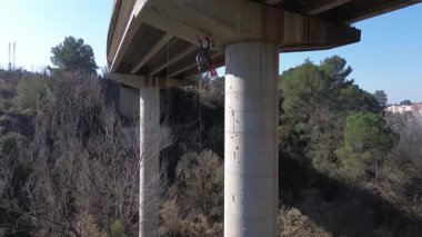 Güvenlik kayışından sarkan halat erişim teknisyeni bakım sırasında otoyol üst geçidinin yapısal bütünlüğünü incelerken beton destek sütunlarını inceliyor.