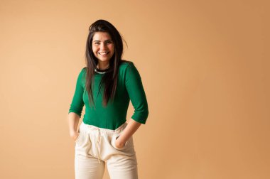 Elleri ceplerinde, günlük giysiler içinde bej bir arka planda gülümseyen neşeli genç bir kadının stüdyo portresi.