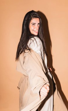 Stüdyo ortamında bej bir arka planda trençkot giyerken ve gülümserken manken.