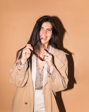 Gözlüğünü tutan ve bej bir arka planda gülen neşeli bir iş kadınının stüdyo portresi.