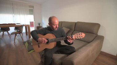 Oturma odasındaki kanepede rahat bir şekilde otururken akustik gitar çalan yetenekli bir müzisyen melodik performans yoluyla müzik tutkusu ifade ediyor.