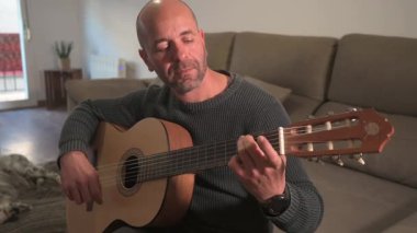Klasik müzik besteleyen yetenekli gitarist oturma odasındaki koltukta parmak stili tekniği uyguluyor, akustik gitar ile melodik ifadeleri keşfediyor.