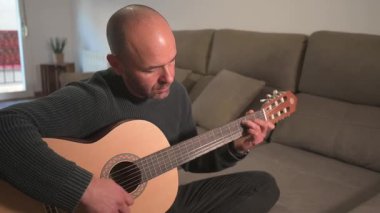 Oturma odasındaki kanepede rahat bir şekilde otururken akustik gitar melodisi çalan yetenekli bir müzisyen, samimi ev performansı yoluyla müzikal tutkusunu ifade ediyor.