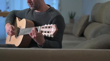 Oturma odasındaki kanepede rahatça otururken melodik akustik gitar çalan bir adam konsantrasyonunu bozmadan yaratıcı müzik tutkusunu ifade ediyor.