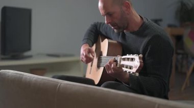 Klasik akustik gitar çalan yetenekli bir gitarist oturma odasındaki koltukta rahatça otururken melodik parmak tarzı performans yoluyla müzik tutkusu ifade ediyor.