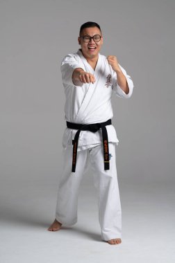 Gülümseyen kyokushin karate dövüşçüsü beyaz karategui ve siyah kuşak giyerek gri arka planda antrenman yaparken yumruk atıyor.