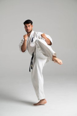 Kyokushinkai karate yapan genç bir adam, parlak bir stüdyo ortamında beyaz keikogi ve siyah kuşak giyerek yükseğe tekme atıyor.