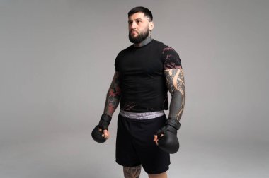 Dövmeli kickboksçu eldiven takıyor, yoğun bir antrenmana hazırlanıyor, stüdyoda gri bir arka plana karşı güven içinde poz veriyor.