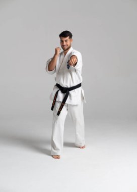 Genç erkek atlet, beyaz üniformalı ve siyah kuşaklı Kyokushinkai karate antrenmanı yapıyor. Tarafsız bir arkaplana güçlü bir yumruk atıyor.