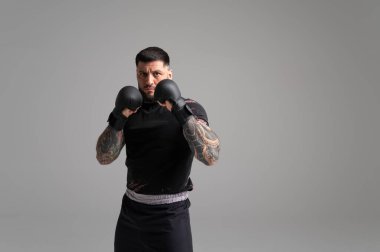 Dövmeli dövüşçü, siyah boks eldiveni giyiyor. Gri bir geçmişe karşı karma dövüş sanatları uyguluyor. Güç ve kararlılık gösteriyor.