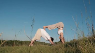 Man and Woman Dressed Alike Doing Difficult Pose While Practicing Yoga Outdoors in the Field with Blue Sky on Background