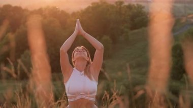 Happy Young Woman with Folded Hands and Closed Eyes Sitting on Mat and Meditating Outdoors at Sunset
