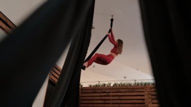 Young woman in a red athletic suit does complex exercises on the canvas, aerial gymnastics in studio