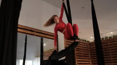 Aerial gymnastics in the studio, young woman in a red athletic suit does complex exercises on canvas