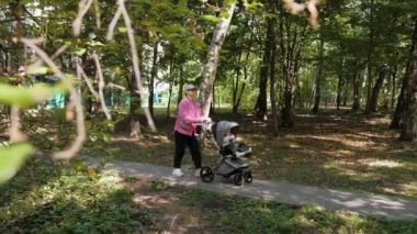 Young stylish mom with baby carriage in the park, mother walking with her little child outdoors