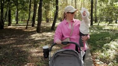 Mother carries her little child in her arms, young stylish mom with a baby carriage in park